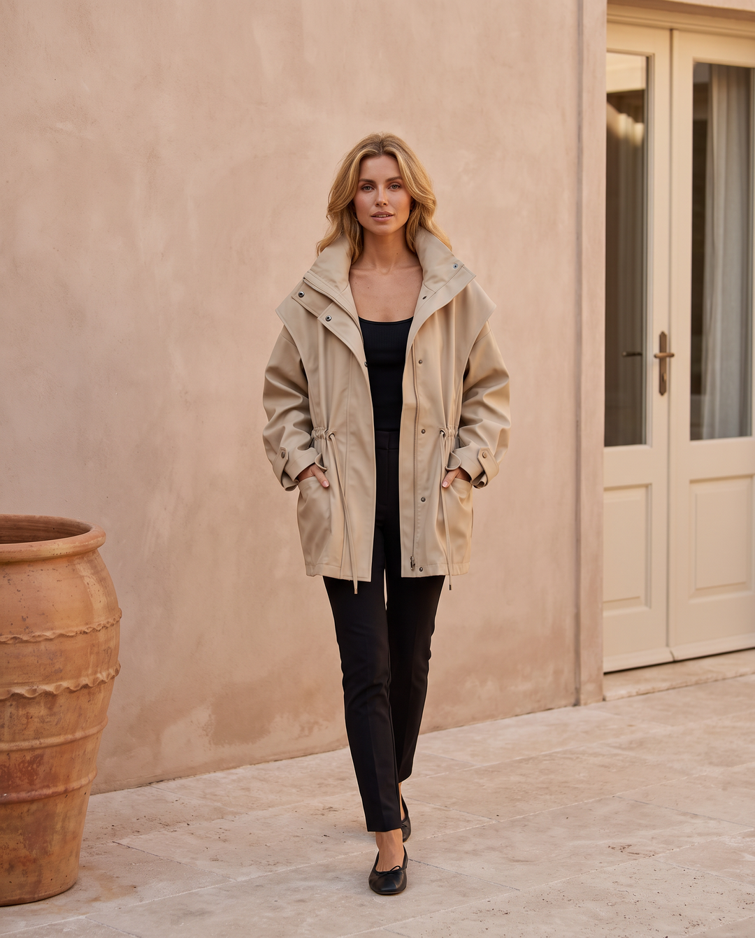 Kovelle beige stand collar windbreaker jacket, on model walking through the Kovelle House courtyard